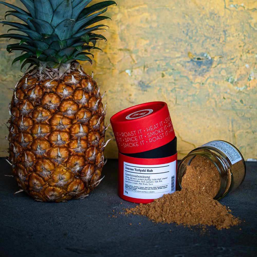 Die Gewürzdose des Ananas Teriyaki Rubs mit dem geöffneten Gewürzglas steht neben einer Ananas vor einer gelb marmorierten Wand