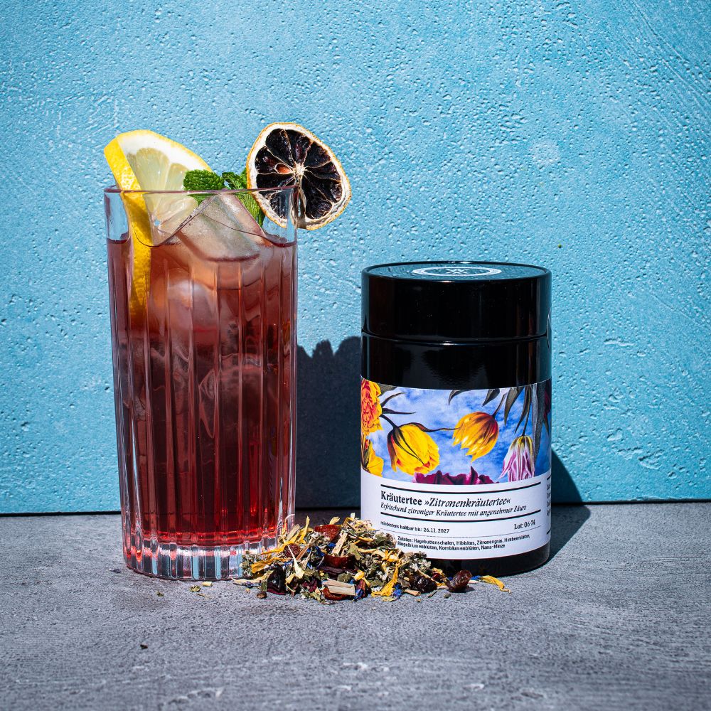 Hohes Glas mit Eistee, dekoriert mit einer Zitronenscheibe neben einer Teedose Zitronenkräutertee und etwas davor ausgeschüttetem losen Tee