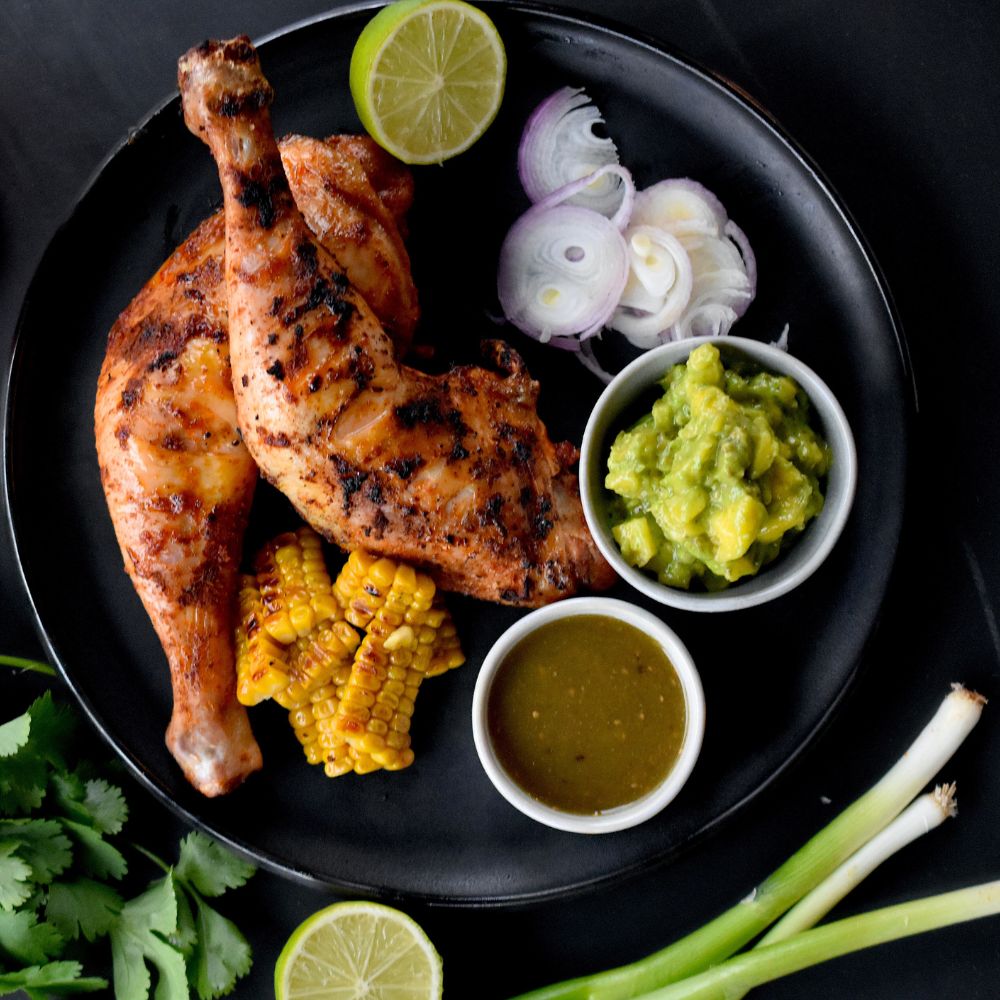 Ein Teller mit zwei Hühnerschenkel mit Jamaican Jerk Chicken glasiert, serviert mit etwas Mais Guacamole und einem Dip.