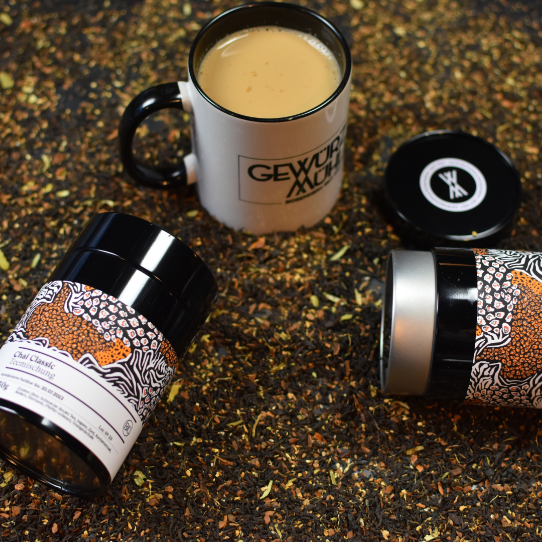 Zwei Chai Classic Gläser liegen auf dem ausgeschütteten Inhalt der Gläser und in der Mitte steht eine Gewürzmühlen-Tasse mit Chai Classic
