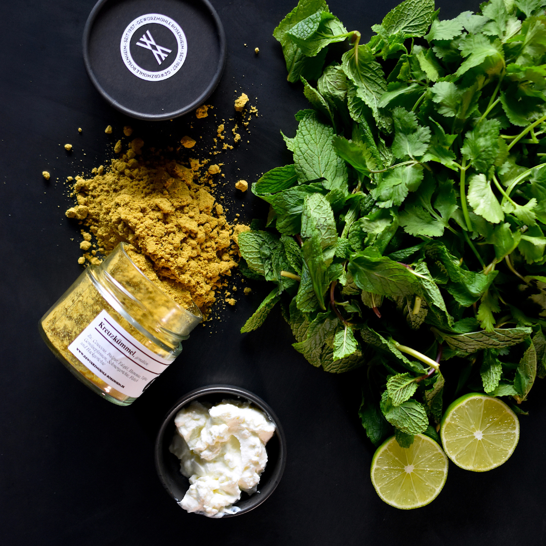 Kreuzkümmel Glas ausgeschüttet auf einem Tisch, daneben liegt Minze und eine aufgeschnittene Limette