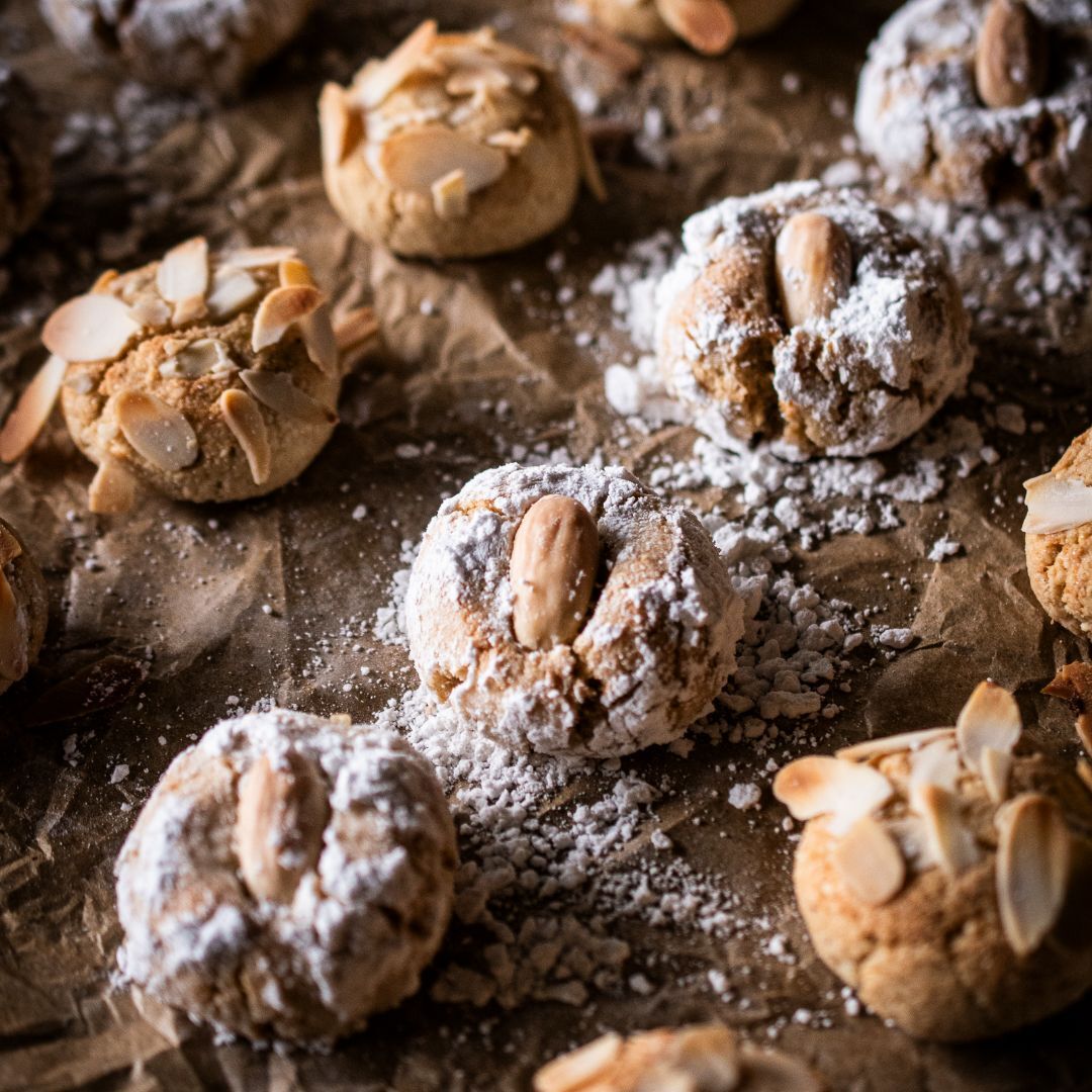 Eine kleine Menge an Amaretti Morbidi mit Keks- & Stollengewürz, die auf Backpapier liegen. Ein Teil der Plätzchen ist mit Mandelplättchen verziert, der andere mit Puderzucker und einer ganzen Mandel.
