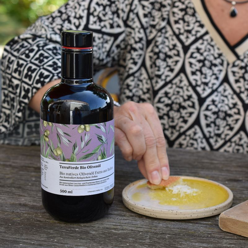 TerraVerde Bio Olivenöl Flasche auf Holztisch, daneben Öl in kleinem Teller, Frau im Hintergrund