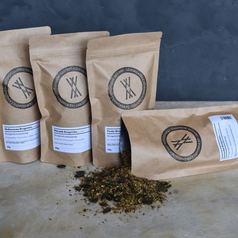 Mehrere braune Papierbeutel mit Gewürzmischungen der Gewürzmühle Rosenheim, davor lose ausgeschüttete Kräuter-Gewürzmischung auf hellem Untergrund