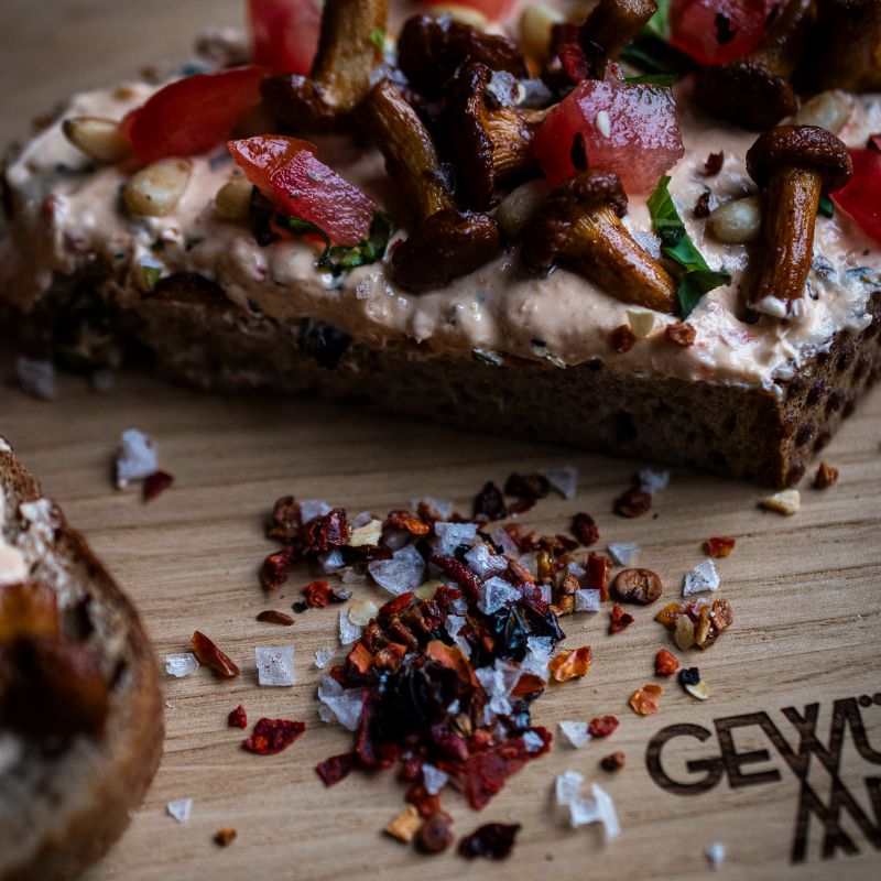 Detailaufnahme des Brotbelags mit cremigem Aufstrich, gebratenen Pfifferlingen, Tomatenwürfeln und Chilisalz-Flocken der Gewürzmühle Rosenheim. Salz- und Chiliflocken liegen auch auf dem Holzbrett mit Gravur des Markenlogos.
