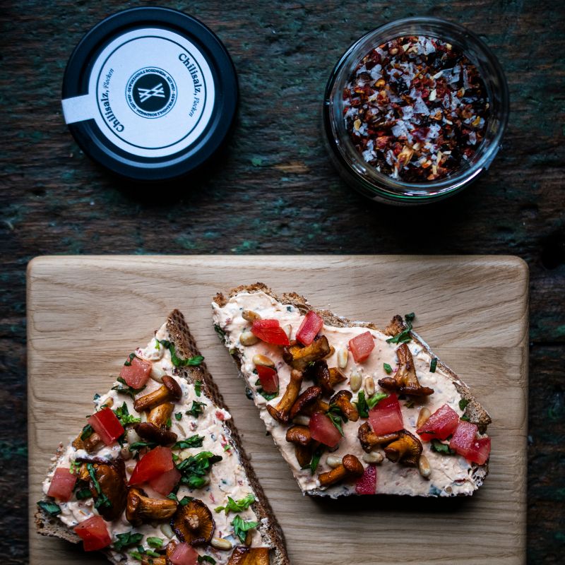 Zwei Brothälften mit Chili-Frischkäse-Creme, Pfifferlingen, Tomaten und Kräutern auf Holzbrett. Daneben ein offenes Glas Chilisalz der Gewürzmühle Rosenheim mit groben roten und weißen Kristallen.