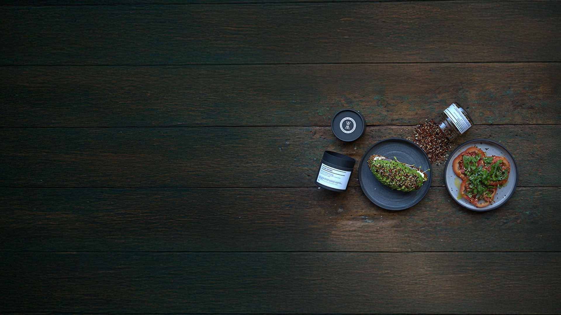 Dunkler Holztisch mit minimalistischer Food-Inszenierung: ein Teller mit Avocadobrot, daneben ein offenes Gewürzglas, ein zweiter Teller mit Tomaten und Basilikum sowie eine schwarze Gewürzdose – stilvolles Foodstyling mit Fokus auf Gewürze und Genuss